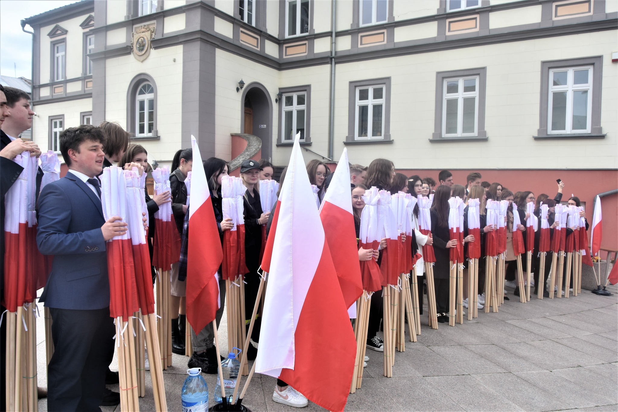 Duża grupa młodzieży z polskimi flagami stojąca na rynku.