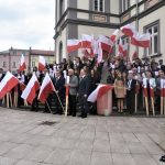 Duża grupa dorosłych i młodzieży z polskimi flagami stojąca na rynku przy ratuszu.