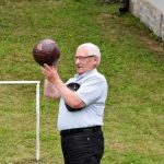 Starszy mężczyzna ubrany w białą koszulę i czarne jeansy, trzyma piłkę w rękach i stoi na murawie boiska.