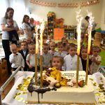 Grupa małych dzieci stoi w jasnej, kolorowej sali i patrzy na tort ze świeczkami i strzelającymi ogniami.