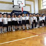 Grupa dzieci chłopców i dziewcząt ubranych w elegnackie odświętne stroje stoi na sali gimnastycznej i spiewa hymn.