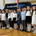 Grupa dzieci chłopców i dziewcząt ubranych w elegnackie odświętne stroje stoi na sali gimnastycznej i spiewa hymn.