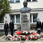 Pomnik z kamienia z półpiersiem Marszałka Józefa Piłsudskiego, przed pomnikiem leżą wiązanki kwiatów, obok pomnika stoją dziewczyny ubrane w zielone mundury. Za nim budynek z jasną elewacją.