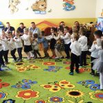 Grupa dzieci dziewczynek i chłopców ubrnaych w odświętne stroje tańczy w sali przedszkolnej na zielonym dywanie w kwiatki kolorowe. Ściany za nimi są kolorowe i wyklejone naklejkami w misie. Występ dzieci ogladają rodzice.