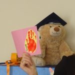 Misiek w czapce absolwenta siedzi na półce, przed nim ręka kobiety trzyma kartkę z odbita kolorową rączką dziecka.