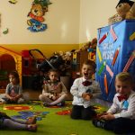 Dzieci chłopcy i dziewczynki ubrane w odświętne stroje siedzą na dywanie koloru zielonego i śpiewają piosenki. Za nimi w koncie poukładane są zabawki.