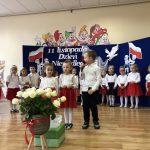 Grupa dzieci odświętnie ubranych stoi w sali przedszkolnej i mówią wierszyki. Na środku stoją dwie dziewczynki i chłopiec, przed nimi bukiet białych róż w wazonie.