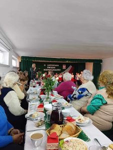 Grupa starszych kobiet siedzi przy stole swiatecznym i smieją się do siebie oraz ogladają występ dzieci.