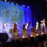 Grupa dziewczynek ubranych na biało siedzi na scenie kina i spiewa kolędy. Za nimi inne dziewczynki również ubrane na biało tańczą.
