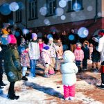 Grupa dzieci ubranych w zimowe kurtki i kombinezony bawią się na płycie rynku. Pada snieg.