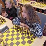 Dziewczynka w długich włosach siedzi przy tablicy szachowej i gra w szachy. Obok niej siedzą inne dzieci.