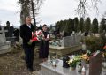 Starszy mężczyzna w garniturze i młoda kobieta stoją przy grobie na cmentarzu. Mężczyzna trzyma w rękach wiązankę kwiatów, zaś kobieta znicz.