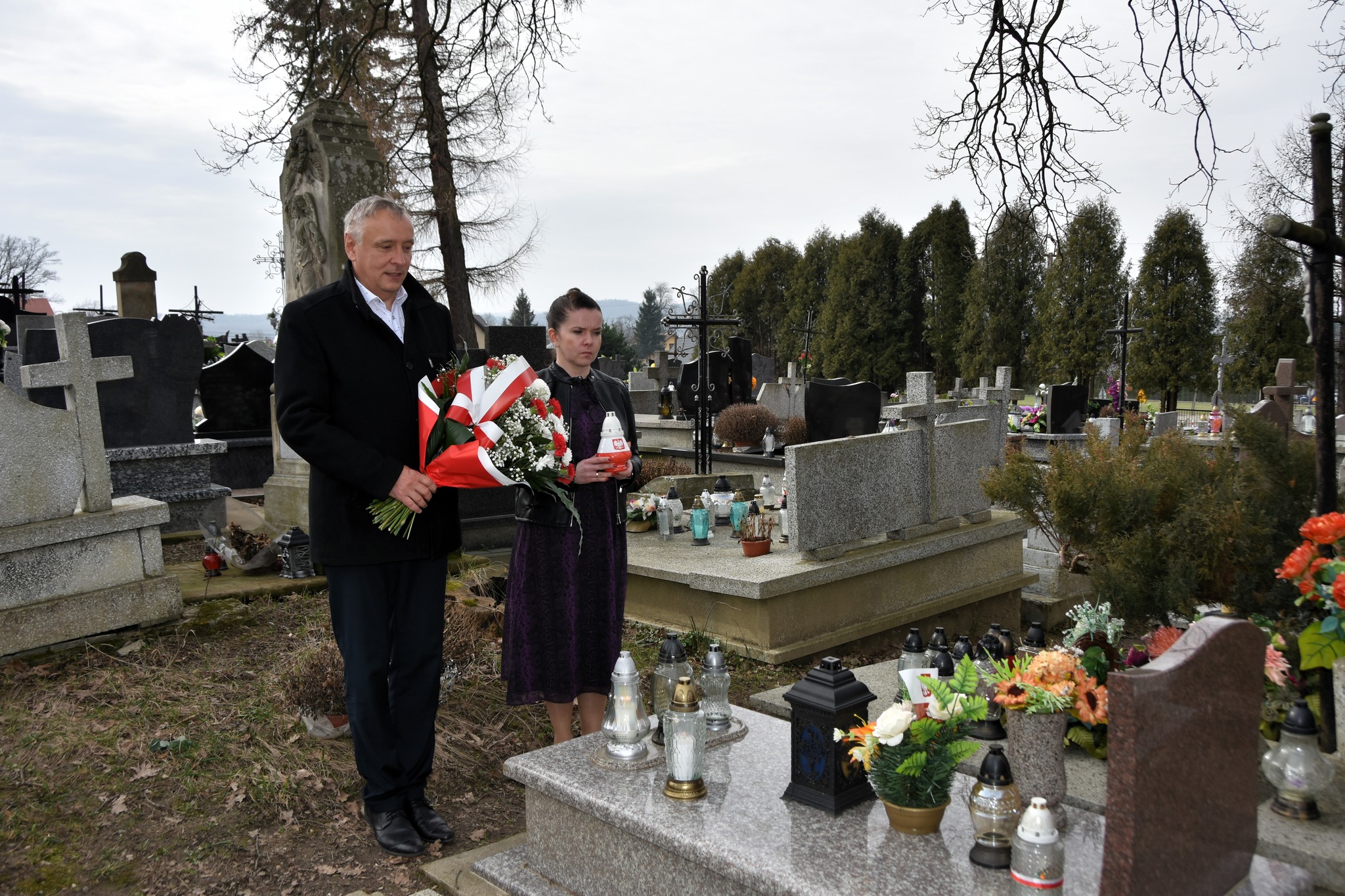 Starszy mężczyzna w garniturze i młoda kobieta stoją przy grobie na cmentarzu. Mężczyzna trzyma w rękach wiązankę kwiatów, zaś kobieta znicz.