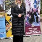 Kobieta w średnim wieku ubrana w czarny długi płaszcz przemawia do mikrofonu na zewnątrz. Za nią stoją ustawione banery promocyjne.